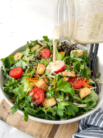 Creamy white balsamic dressing being poured onto a vibrant leafy green salad with tomatoes.